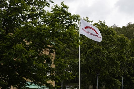 A white Migrationsverket flag flying outside against green trees.