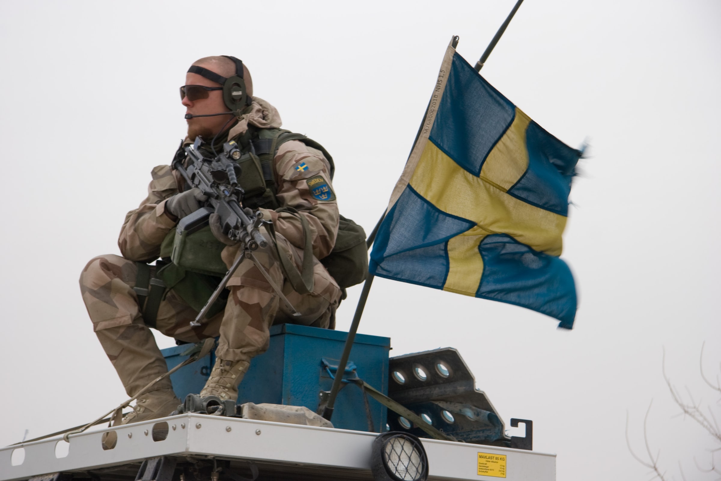 A Swedish soldier in field uniform sits on a military vehicle beside a Swedish flag.