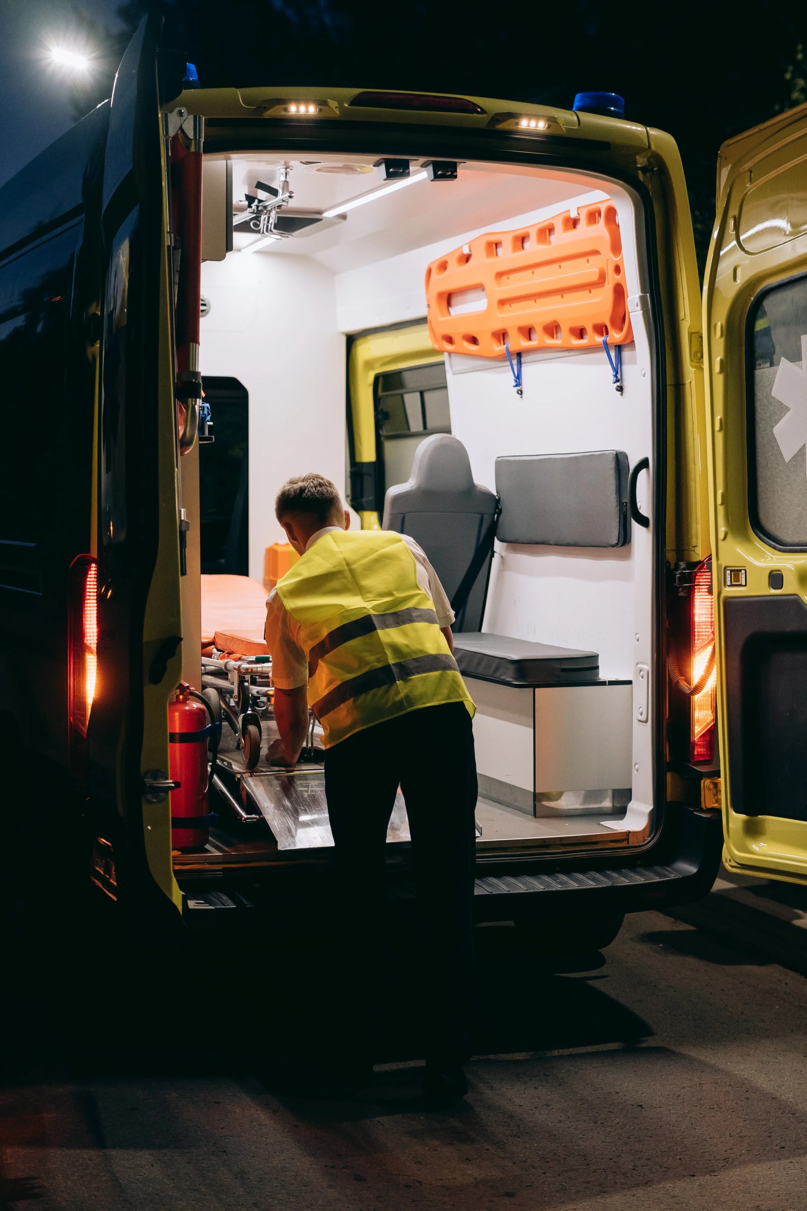 Worker in reflective vest handling a gurney in an open ambulance at night.