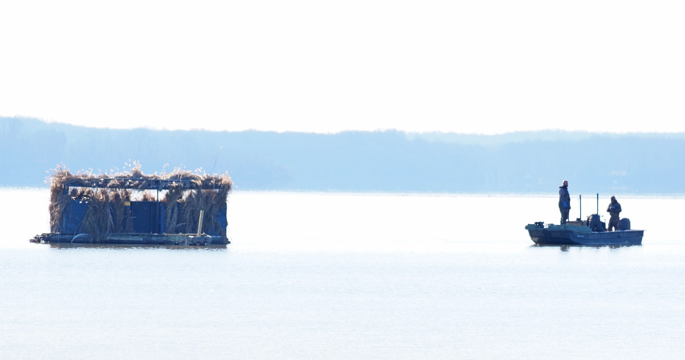 Two duck hunters travel in a small boat beside a reed hunting blind on a lake.