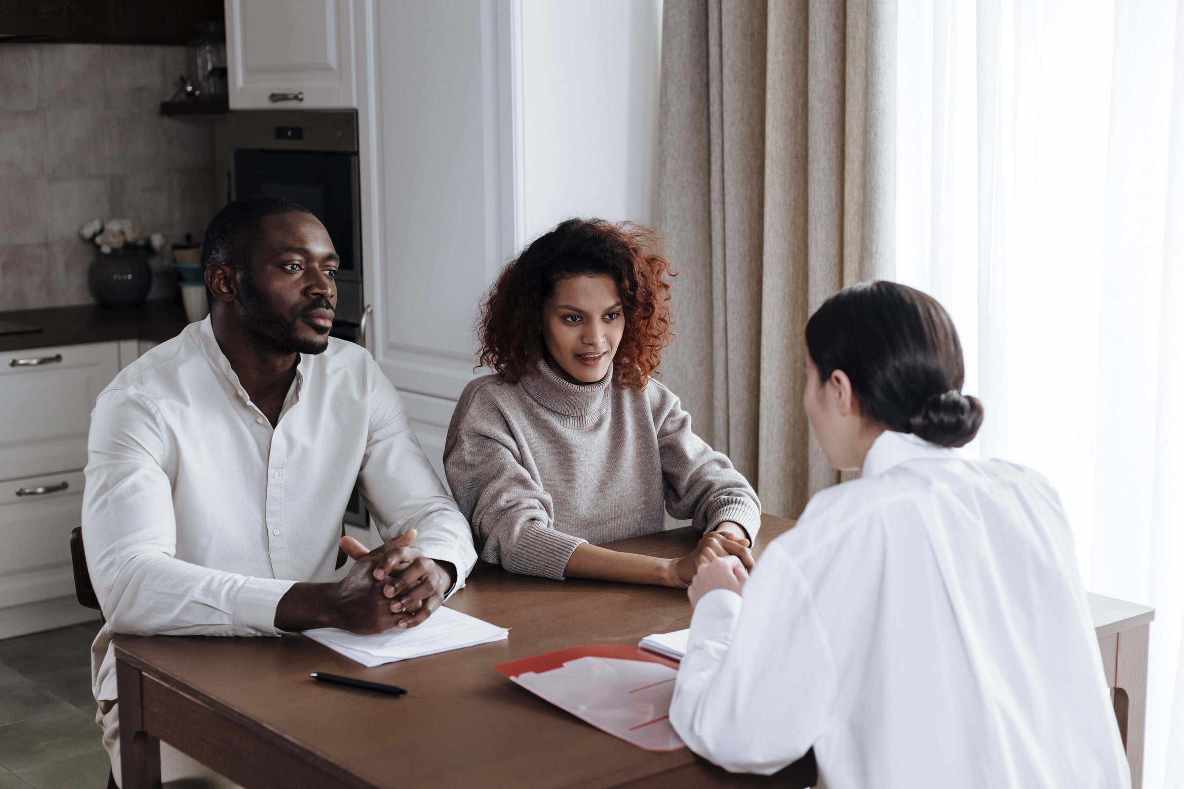A social worker with a red folder speaks with two adults across a table, with papers in front of them.