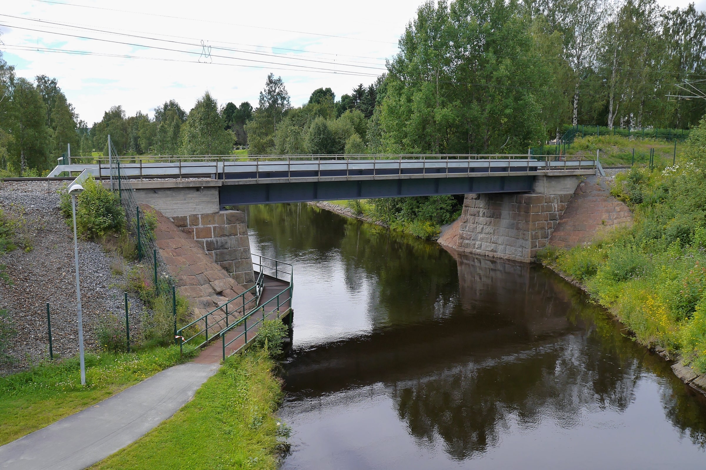 A railway bridge crosses Bodån in central Boden, with a footpath and green trees beside the water.