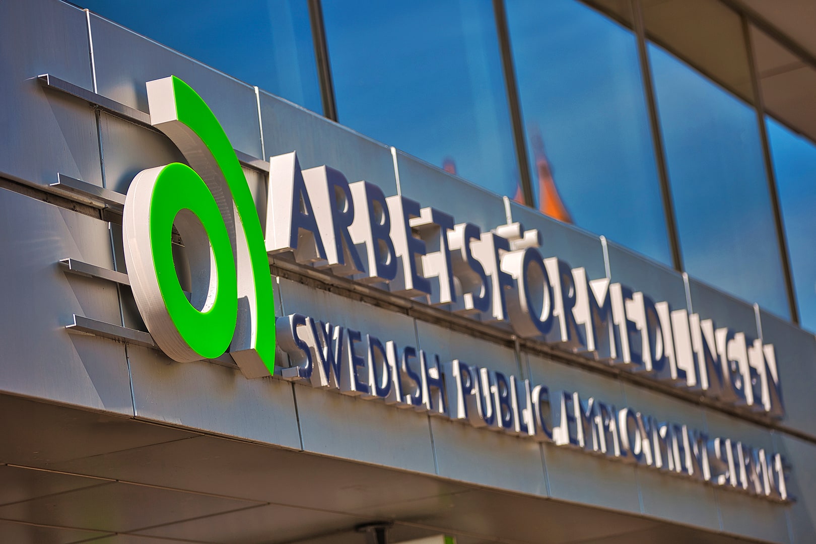 The green Arbetsförmedlingen symbol and bilingual agency name on a glass-fronted building in daylight.