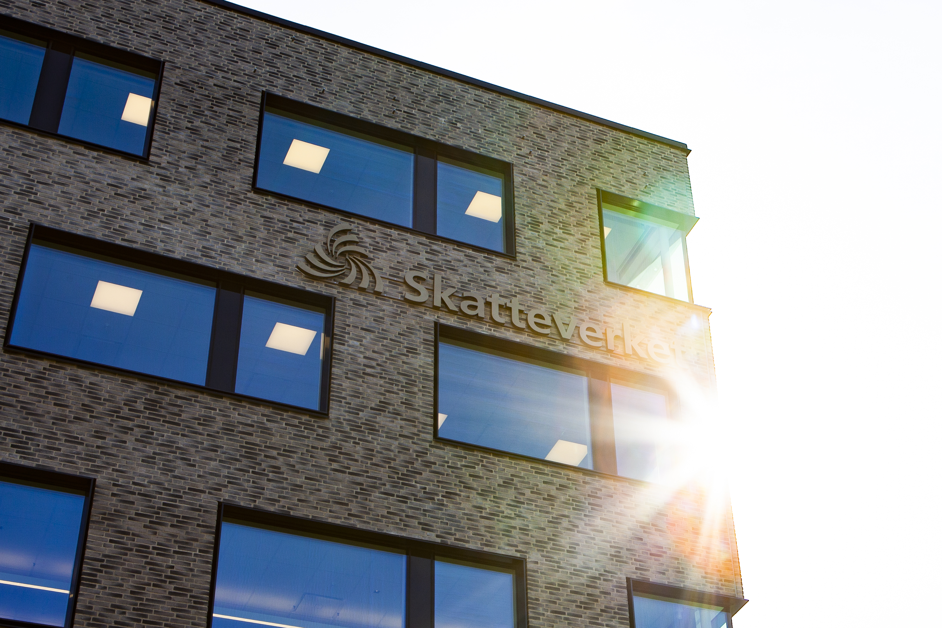 The Skatteverket name and symbol mounted on the brick facade of the Kronan office in Sundbyberg, lit by low sunlight.