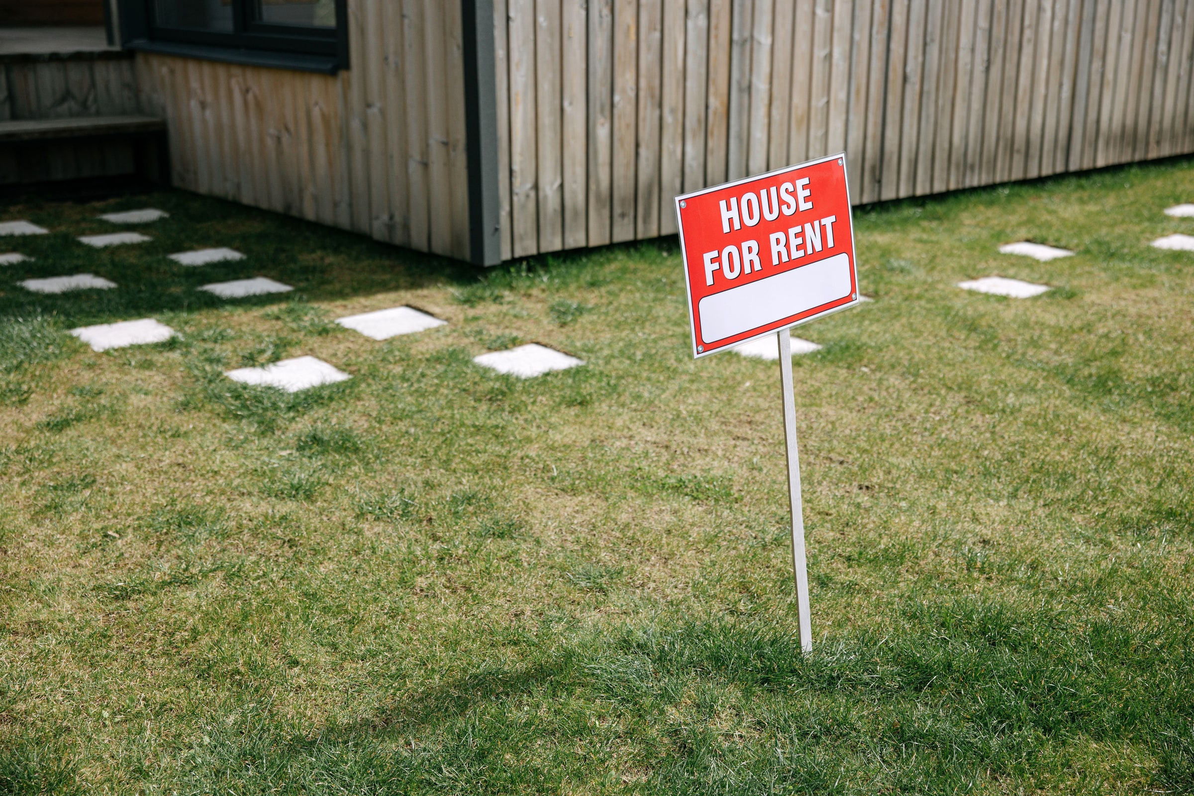 Red and white HOUSE FOR RENT sign on a lawn beside a wooden house.