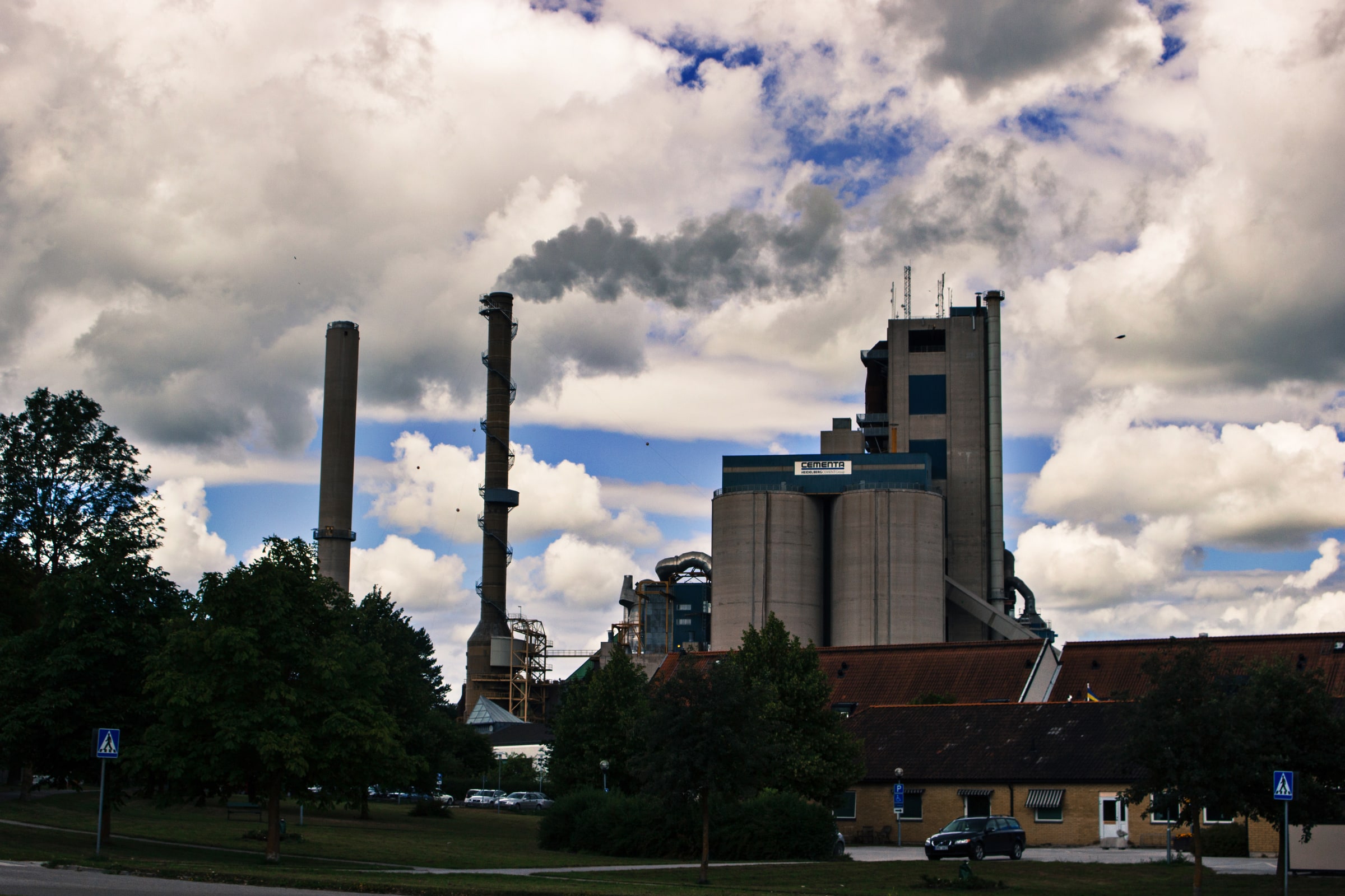 The Cementa cement plant in Slite rises behind trees, with industrial stacks and a visible plume under a cloudy sky.