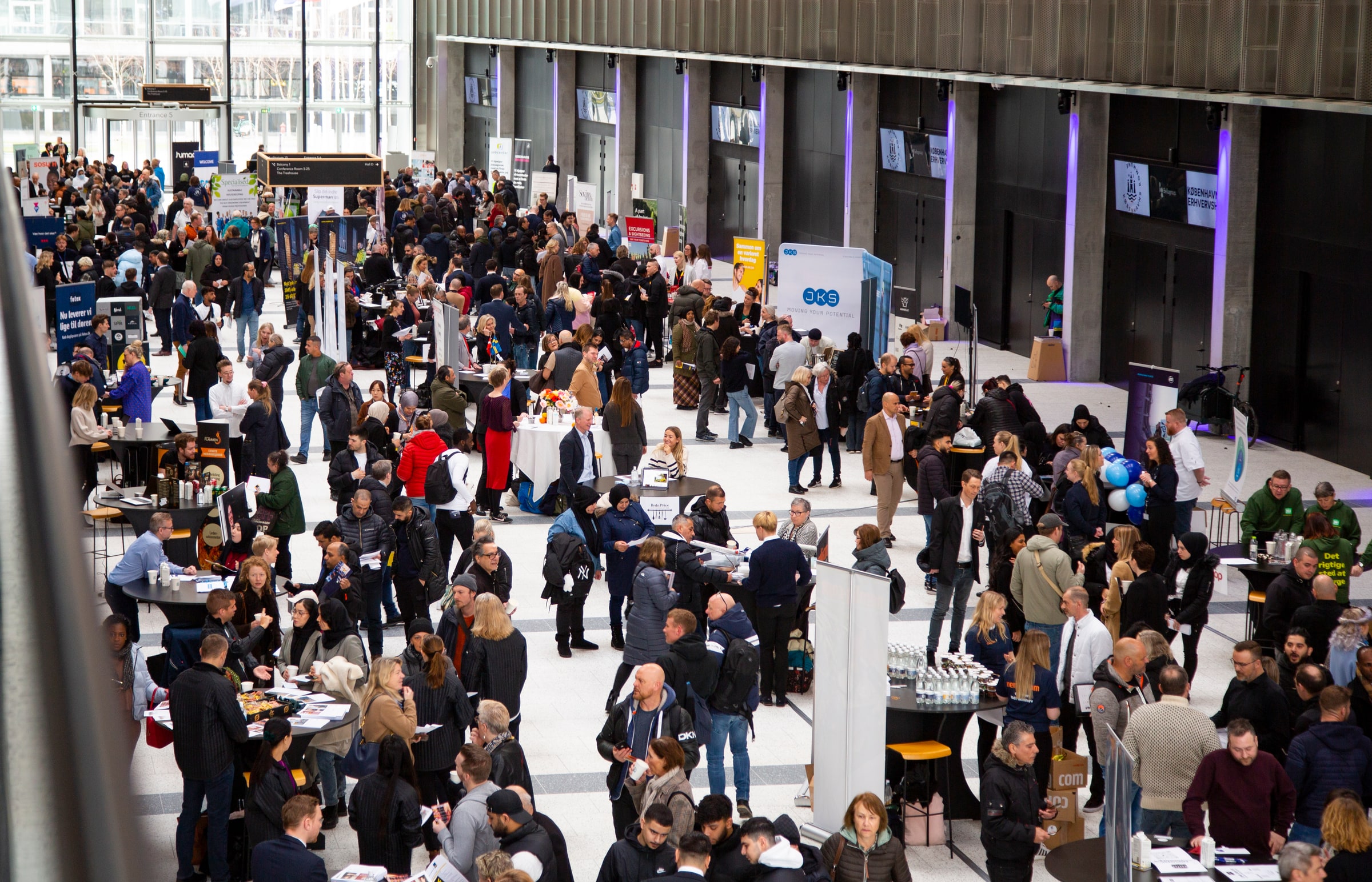 A wide view of a crowded Danish-Swedish job fair with jobseekers and recruiters speaking at many stands.